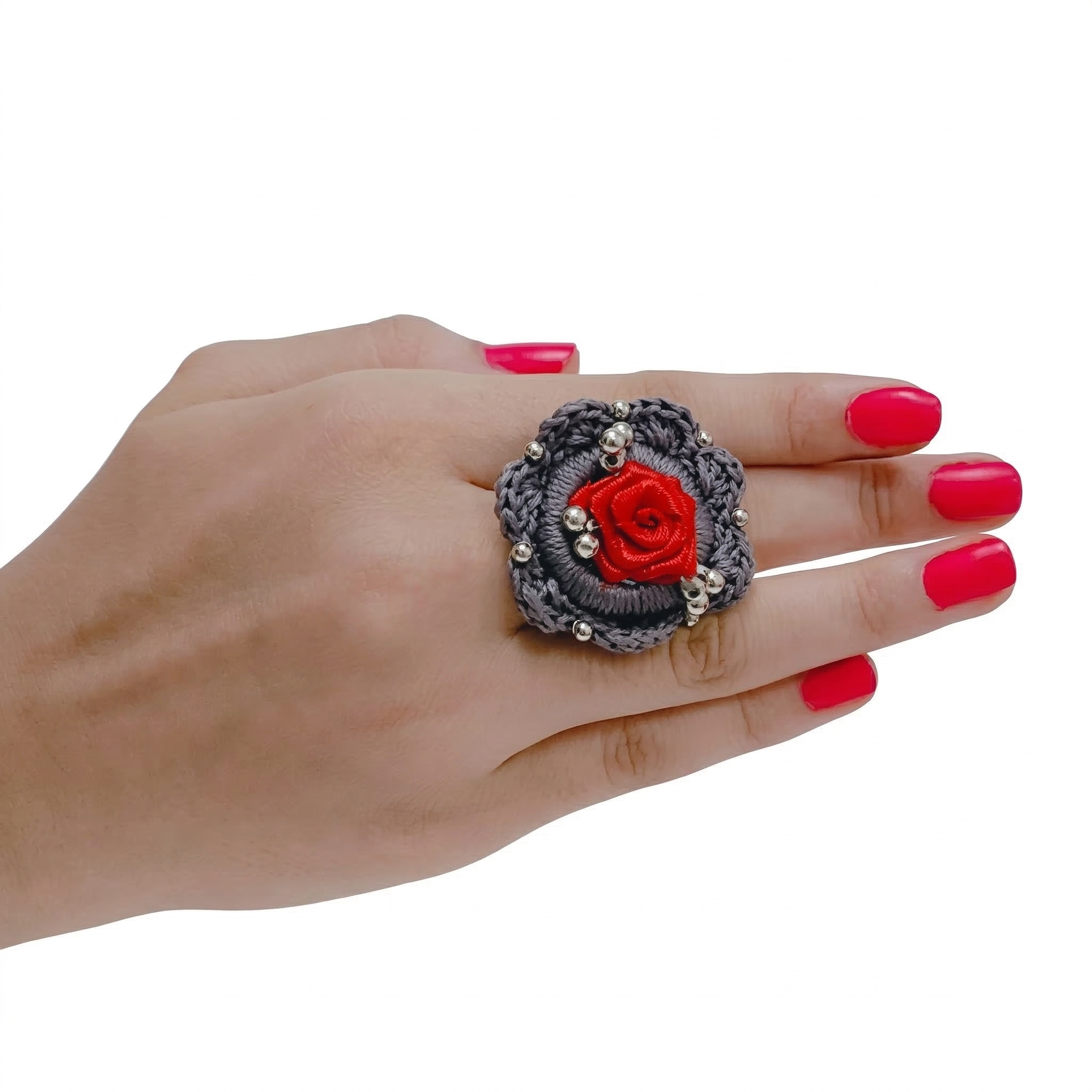 Top view of a grey crochet flower ring with a red fabric rose center and silver bead accents on a model's hand.