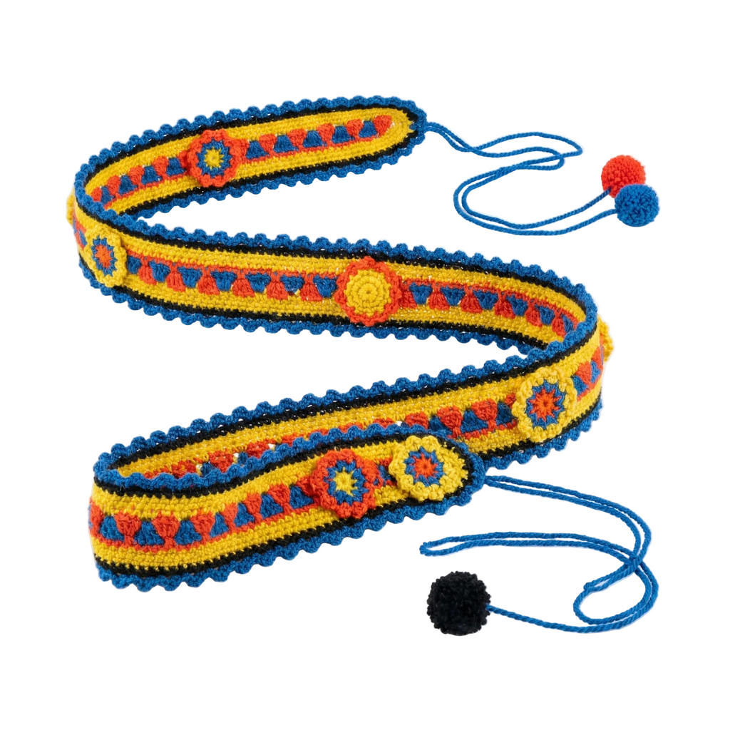 A handcrafted crochet belt coiled on a white background, featuring a bright yellow base, red and blue geometric patterns, decorative floral medallions, a blue scalloped edge, and string ties ending in red, blue, and black pom-poms.