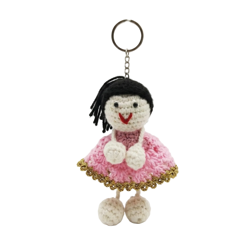 A photorealistic view of a structured cream-colored Saffiano leather satchel handbag with tan top handles and black leather side trim with gold buckles. A handcrafted amigurumi crochet doll charm with black hair, a pink dress, and a smile is attached to the handle ring. The bag sits on a clean, seamless white background.