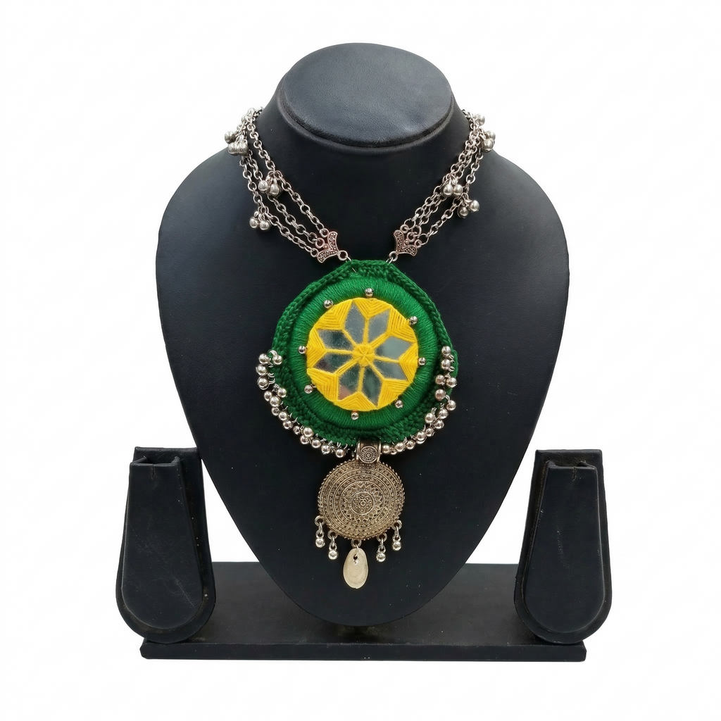 Green fabric mandala necklace with yellow mirror work and oxidized silver charms on a black display stand.
