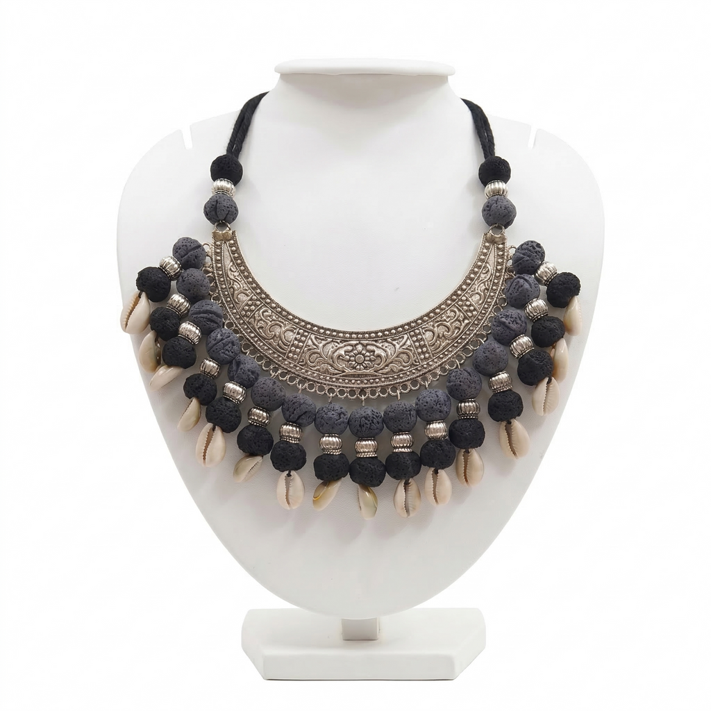Handcrafted tribal statement necklace on a white display stand featuring a large oxidized silver crescent pendant, textured black beads, and dangling cowrie shells.