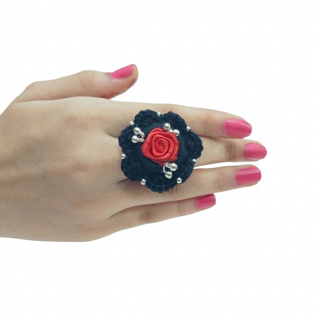 A handmade black crochet cocktail ring featuring a central red satin rose and silver bead accents on a model’s hand.