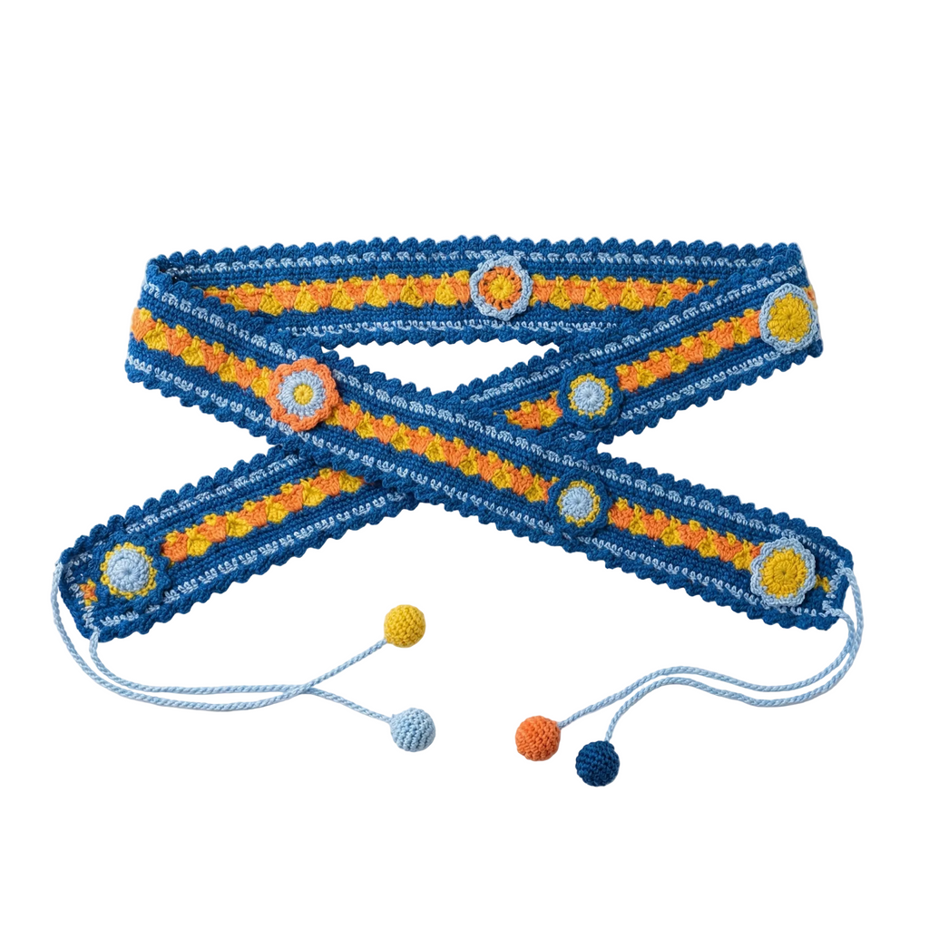 A flat lay photograph of a handmade blue crochet belt featuring orange and yellow floral motifs and light blue string ties with colorful pompoms on a white background.