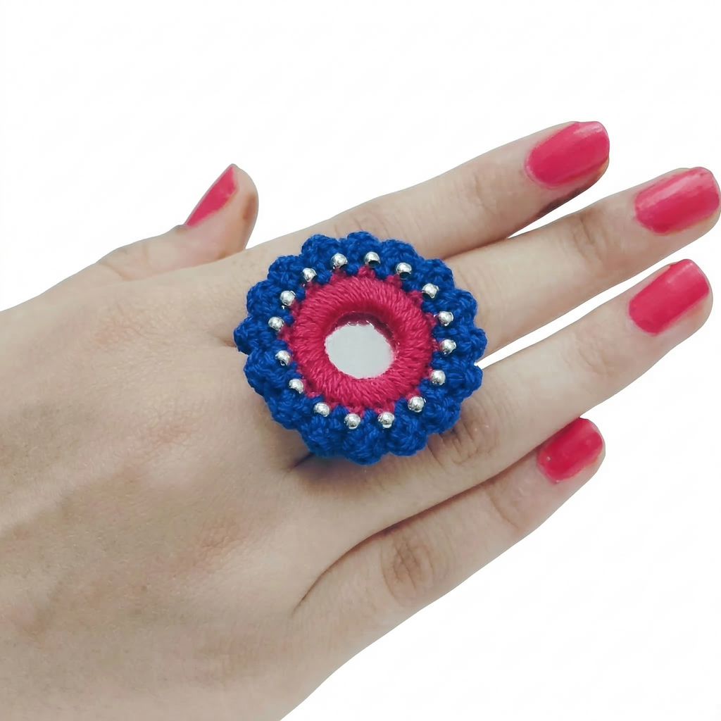 A handcrafted blue and pink crochet statement ring featuring a central round mirror and silver bead accents on a white background.