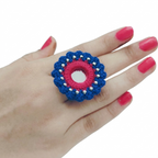 A handcrafted blue and pink crochet statement ring featuring a central round mirror and silver bead accents on a white background.