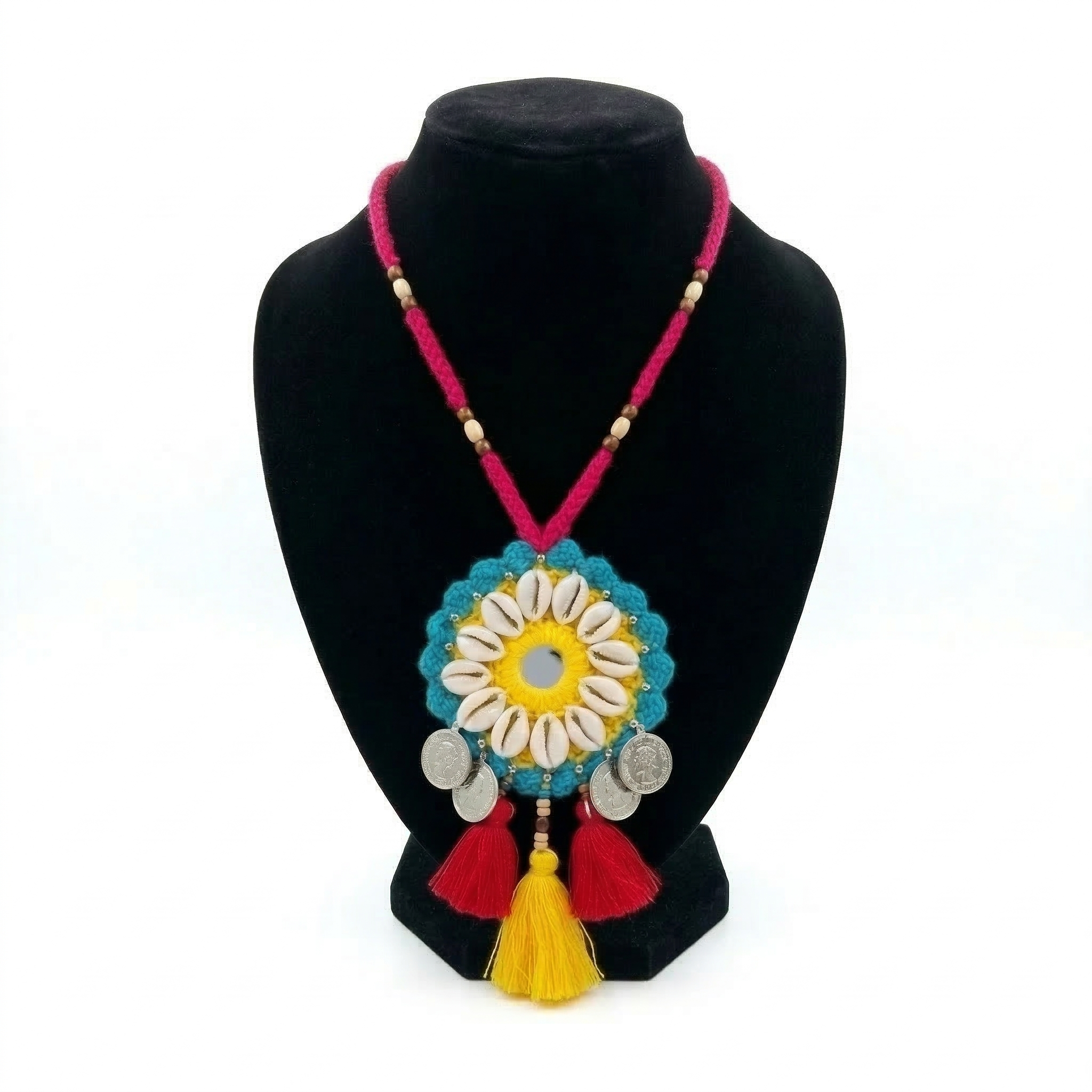 Traditional Indian handmade crochet pendant necklace featuring white cowrie shells, silver coins, and vibrant red and yellow tassels on a black display stand.