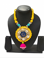 Handmade yellow thread necklace on a black display stand, featuring a circular pendant with white cowrie shells, blue mirror work, and a pink jhumka tassel.