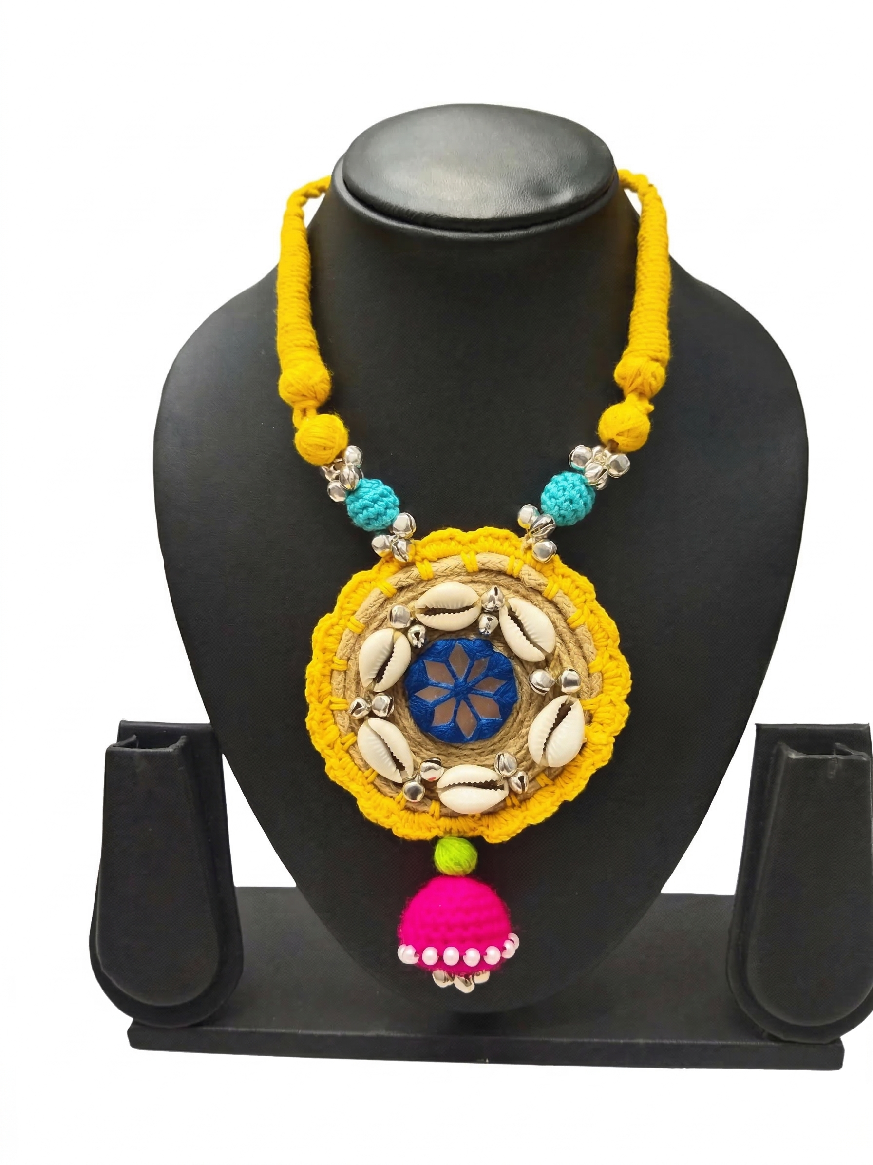 Handmade yellow thread necklace on a black display stand, featuring a circular pendant with white cowrie shells, blue mirror work, and a pink jhumka tassel.