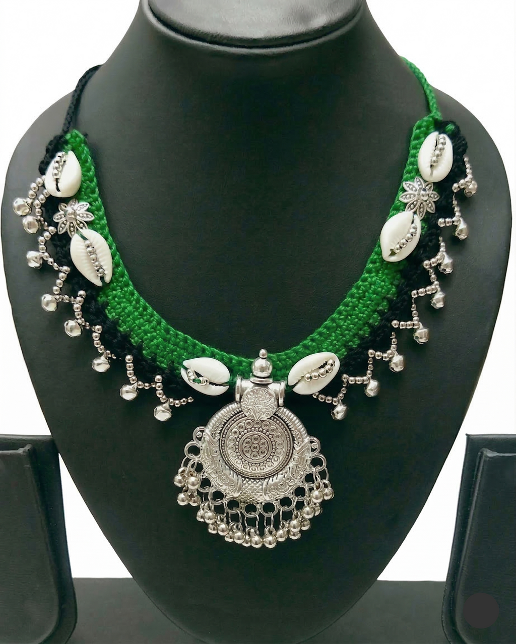Green and black crochet necklace featuring white cowrie shells and a circular silver-toned oxidized metal pendant with dangling beads on a display stand.