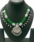 Green and black crochet necklace featuring white cowrie shells and a circular silver-toned oxidized metal pendant with dangling beads on a display stand.