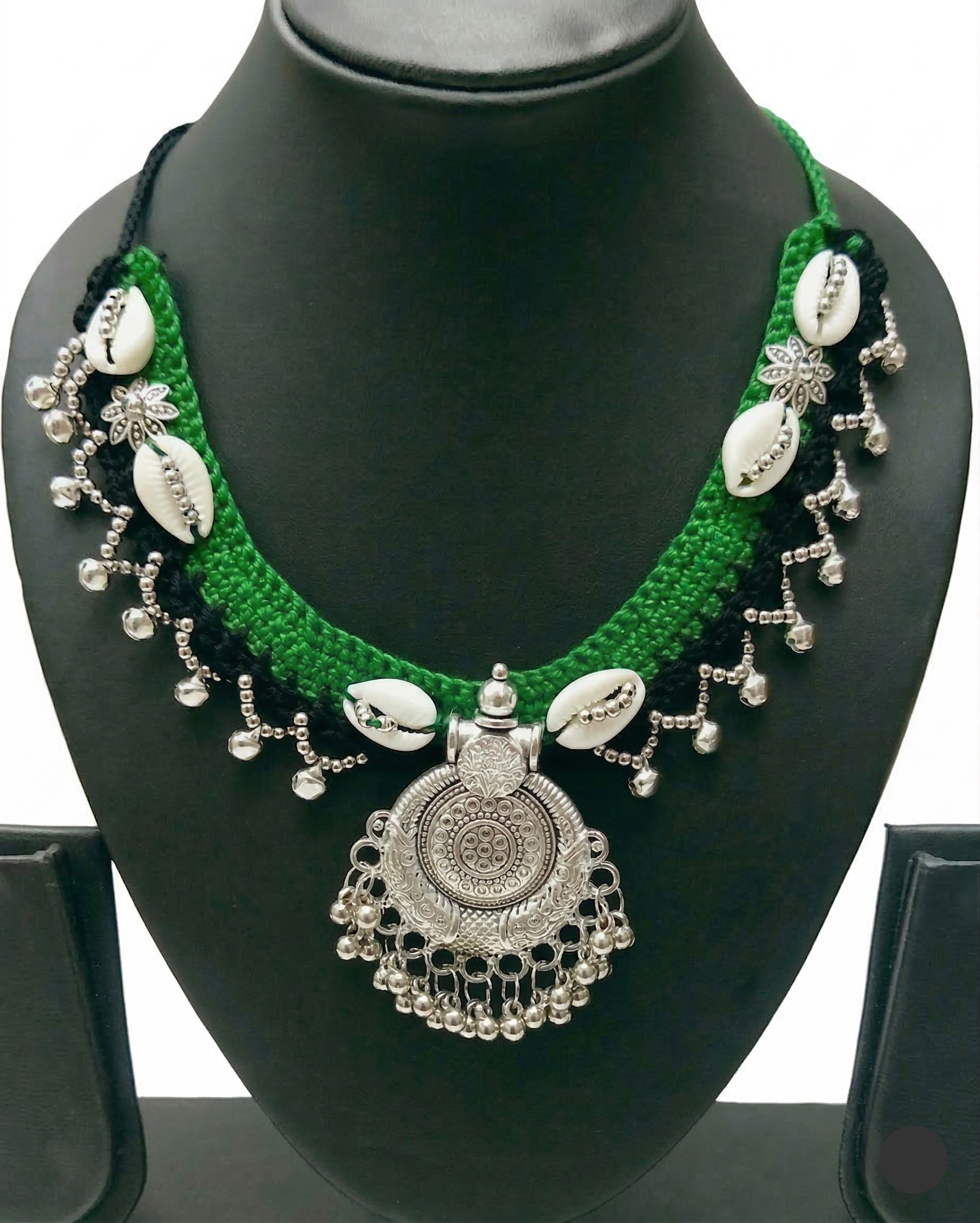 Green and black crochet necklace featuring white cowrie shells and a circular silver-toned oxidized metal pendant with dangling beads on a display stand.