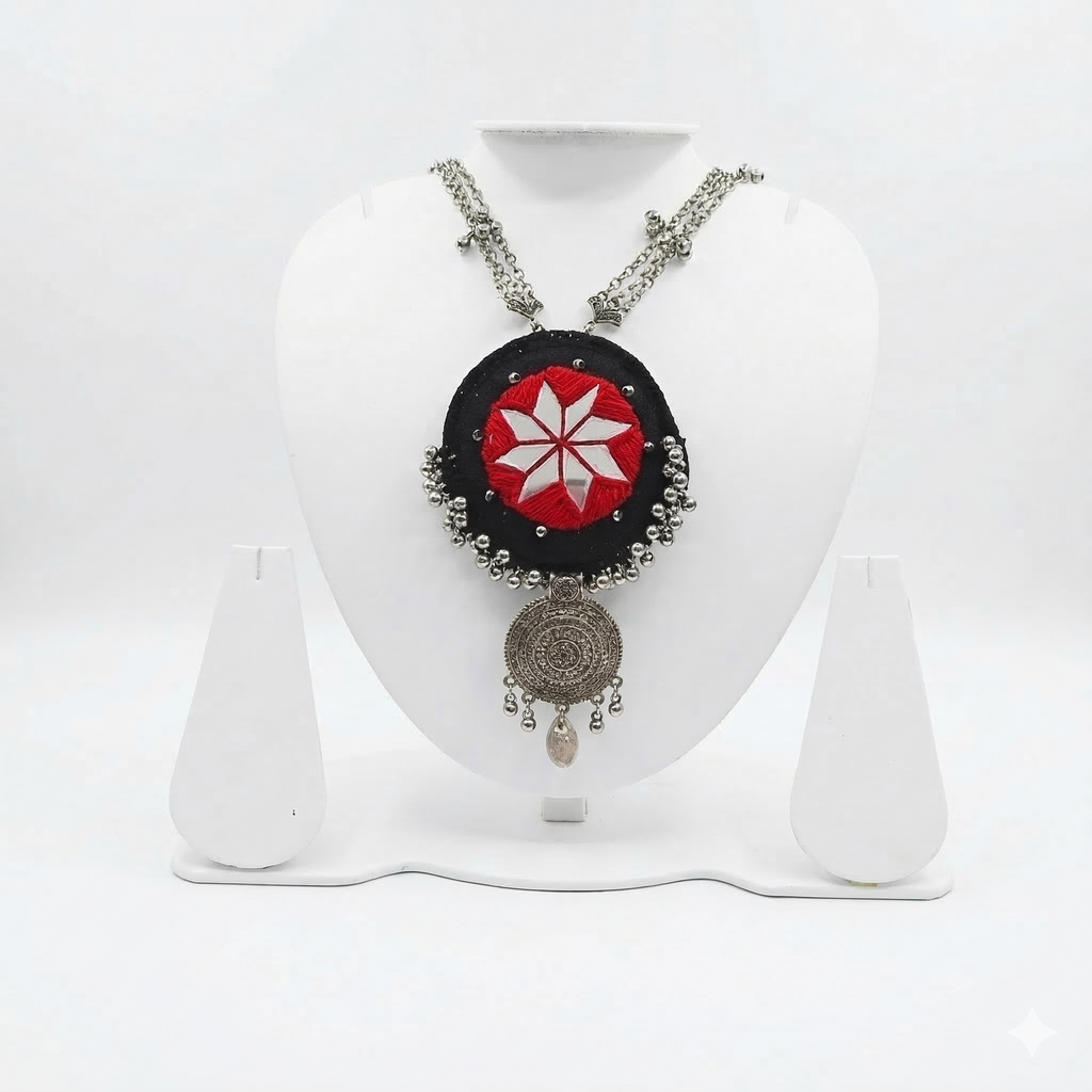 Square product shot of an oxidized silver tribal necklace with a red and black mirror-work pendant on a white jewelry stand.