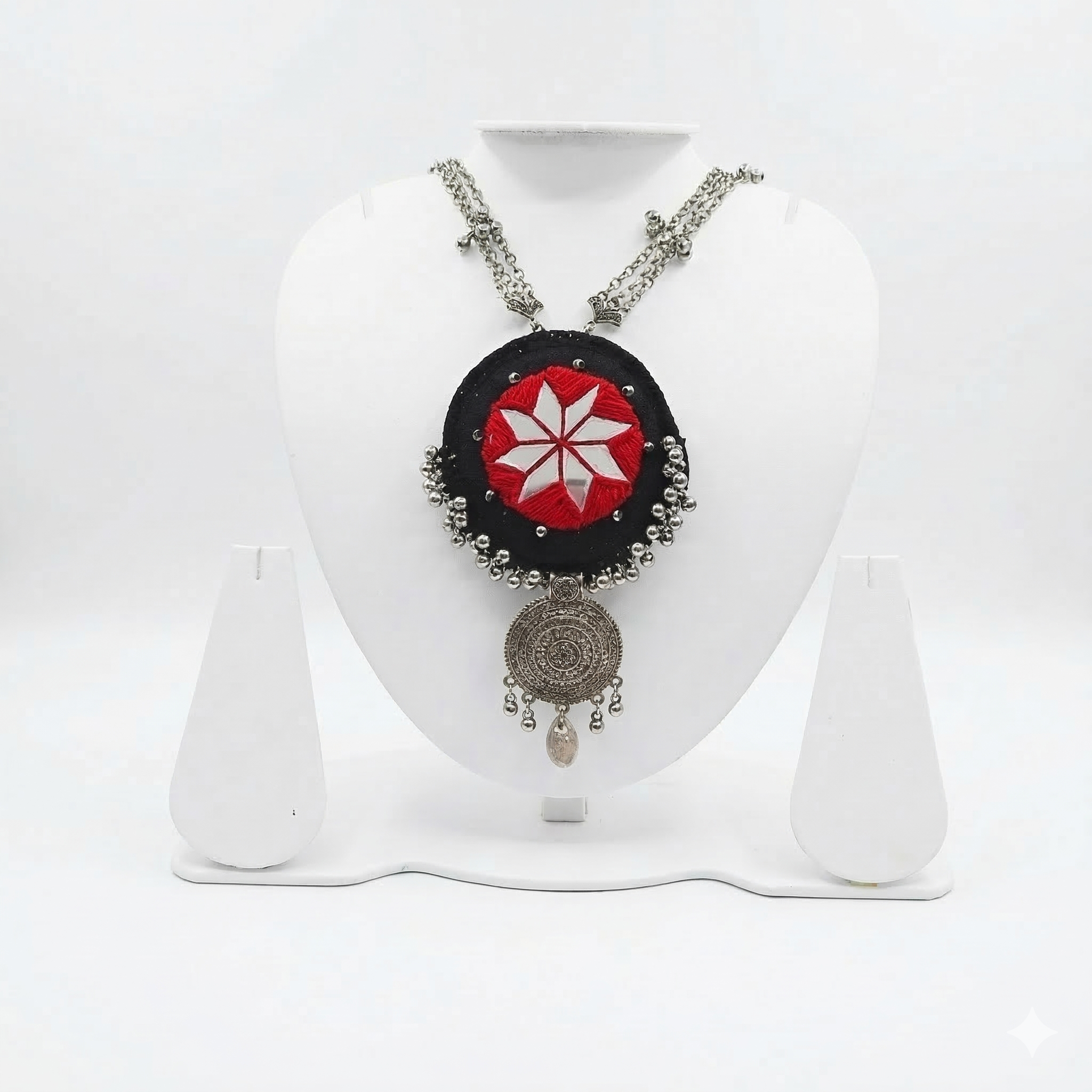 Square product shot of an oxidized silver tribal necklace with a red and black mirror-work pendant on a white jewelry stand.