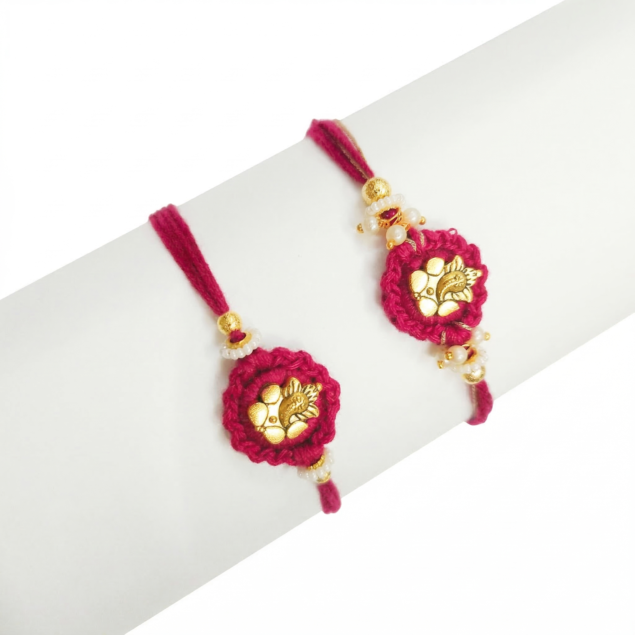 Two handcrafted magenta pink rakhis featuring gold-toned Ganesha motifs and white pearl beads on a white display stand.