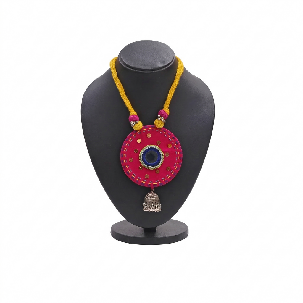 Pink fabric medallion necklace with mirror work, yellow braided cord, and silver jhumka charm on a black display stand.