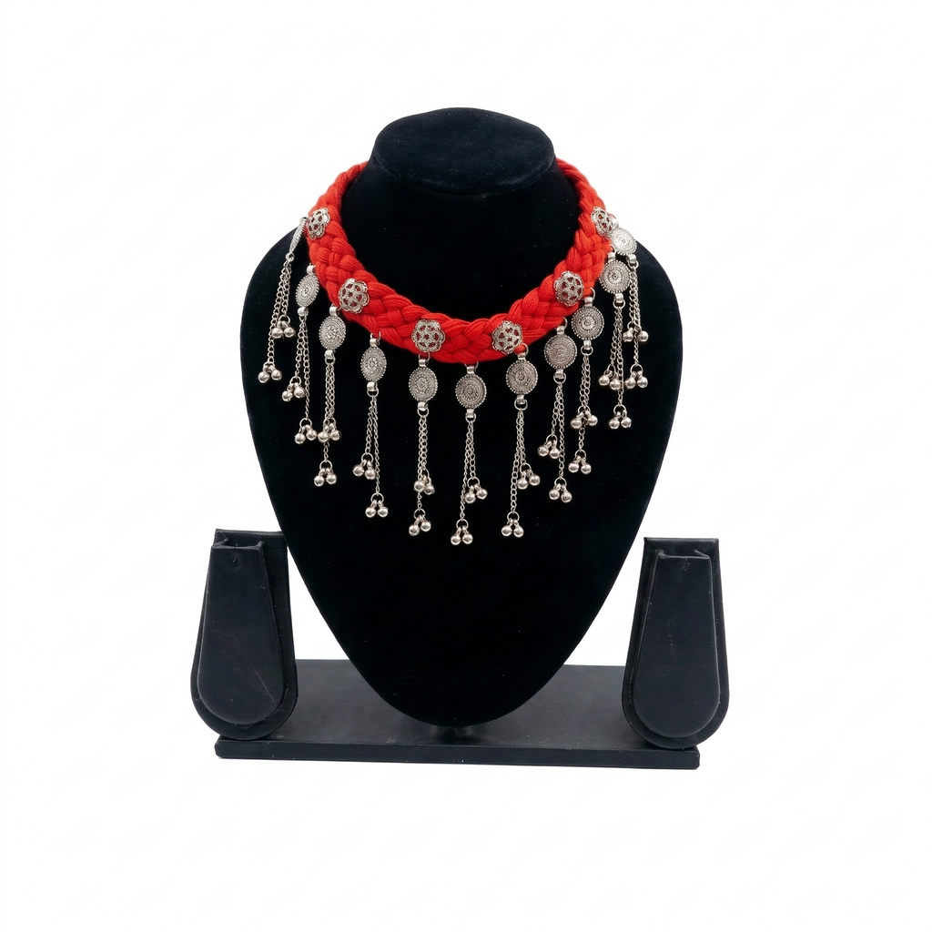 Red braided fabric choker necklace on a black display stand featuring hanging oxidized silver coins and ghungroo bells.