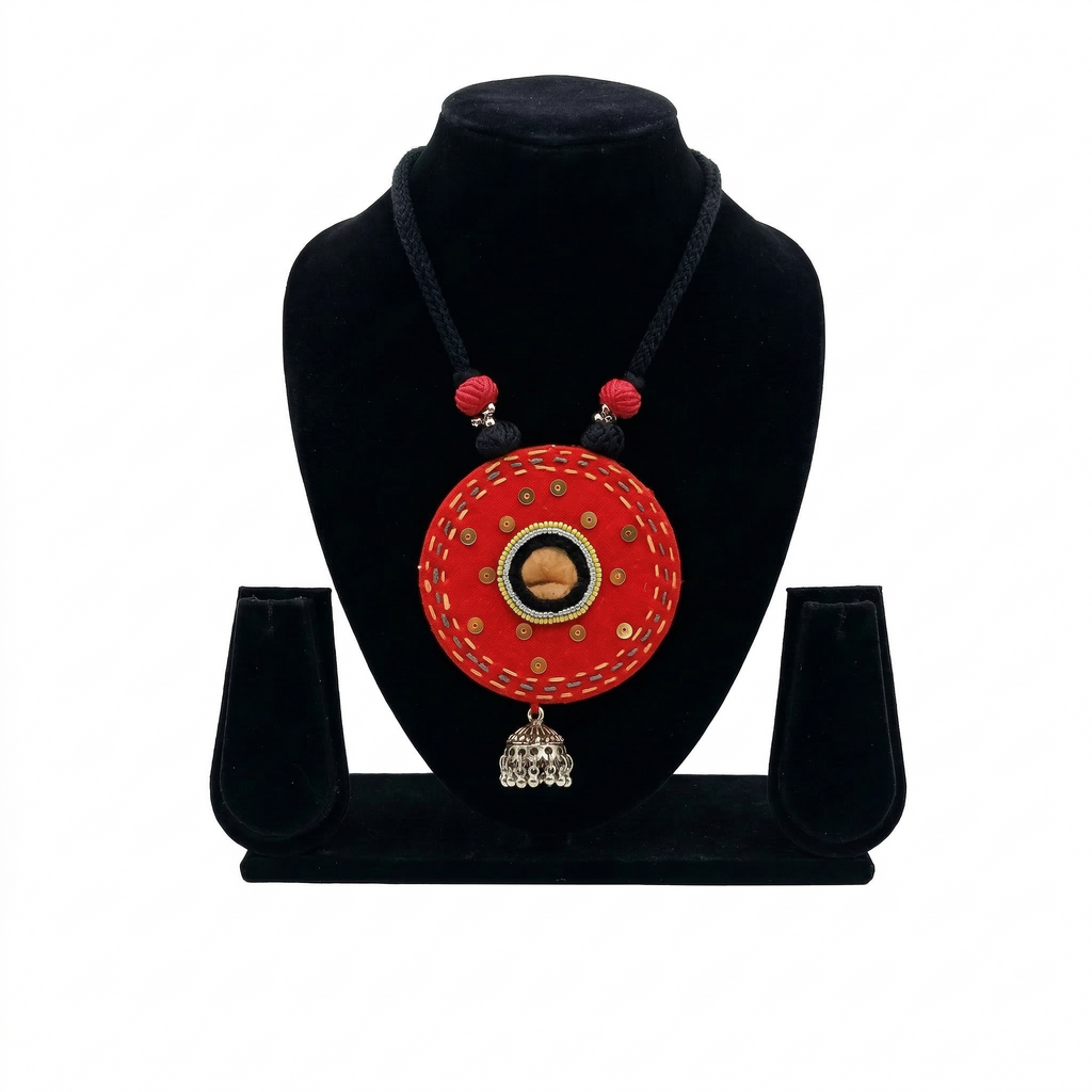 Handcrafted circular red fabric necklace with mirror work detailing and a silver metal jhumka bell pendant on a black bust.