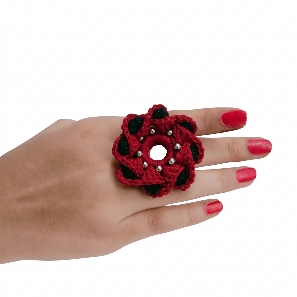 A handcrafted red and black crochet flower ring with silver bead details worn on a hand with pink nail polish, isolated on a white background.