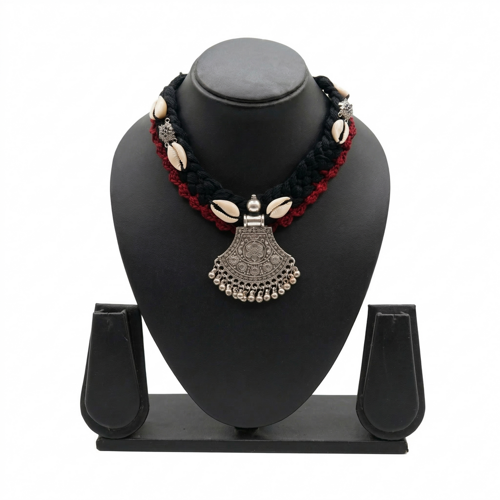 Black and red braided fabric necklace featuring white cowrie shells and a traditional oxidized silver-finish tribal pendant on a black display stand.