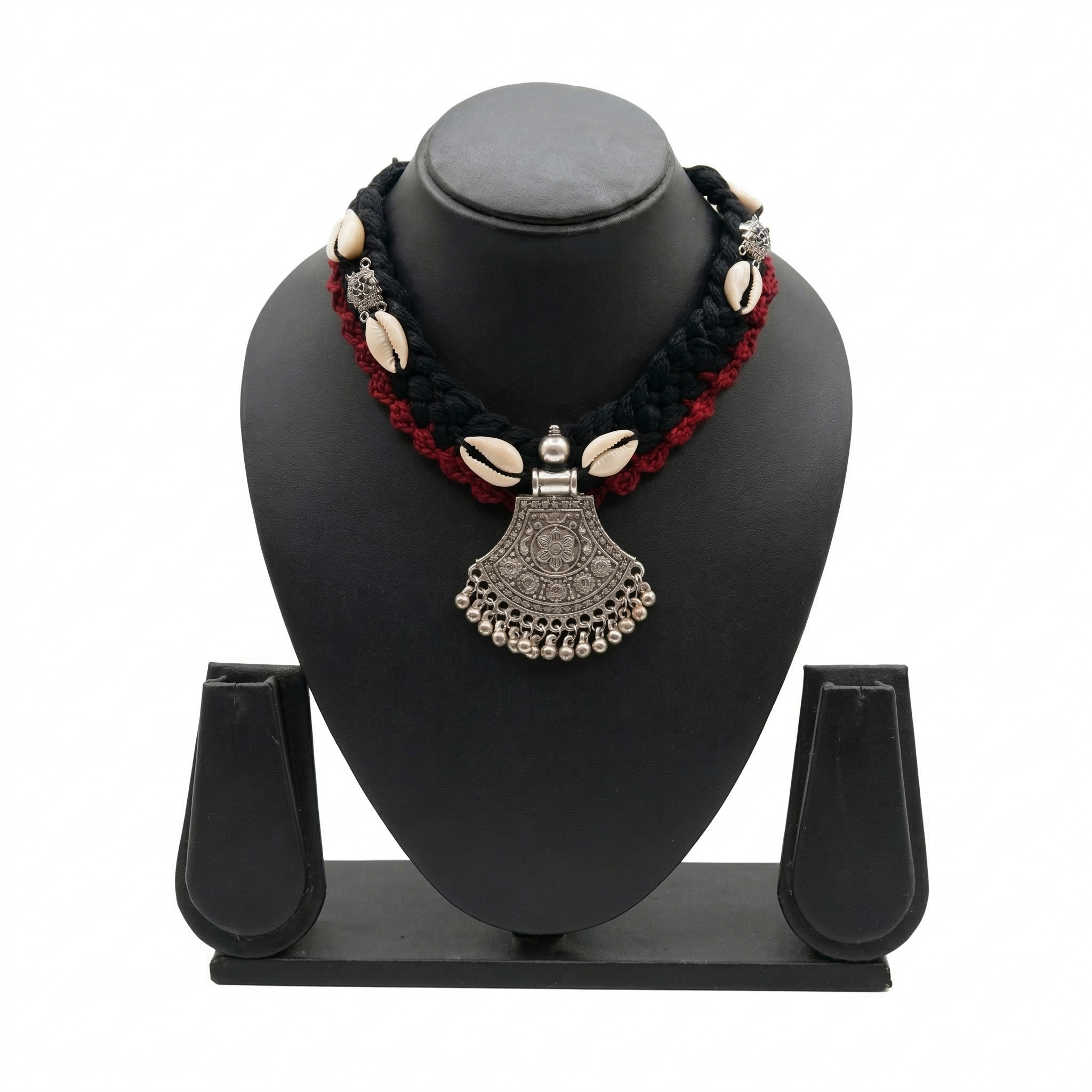 Black and red braided fabric necklace featuring white cowrie shells and a traditional oxidized silver-finish tribal pendant on a black display stand.
