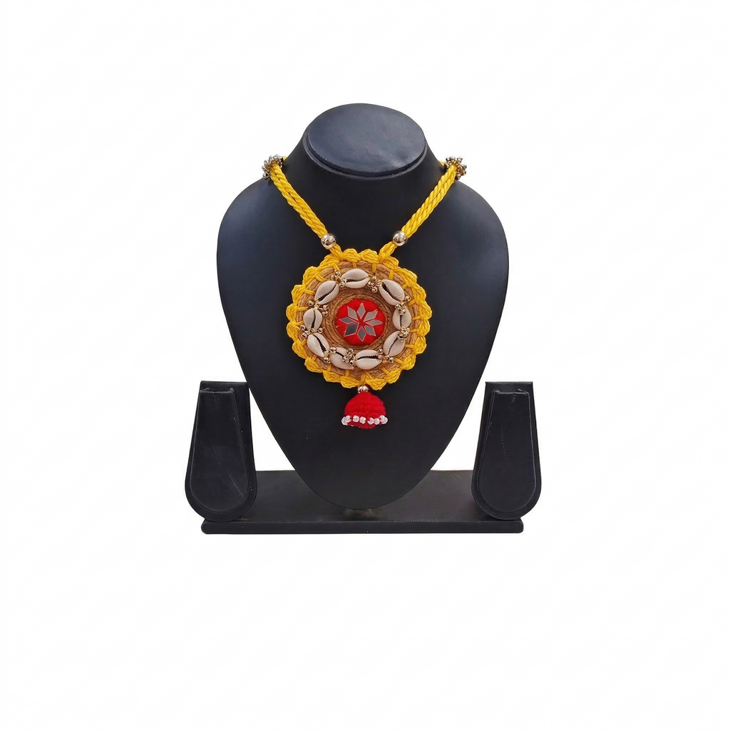 Yellow braided cord necklace featuring a circular pendant with cowrie shells, red mirror work center, and red tassel on a black display stand.