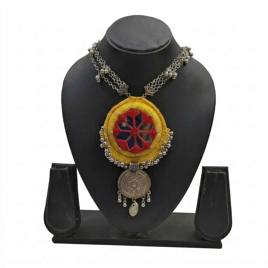 Handmade yellow fabric necklace with red mirror work embroidery and silver oxidized mandala pendant on a black display stand.