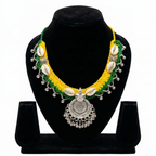 Vibrant yellow and green crochet necklace on a black bust featuring white cowrie shells, silver flower accents, and a large circular oxidized silver pendant with dangling beads.