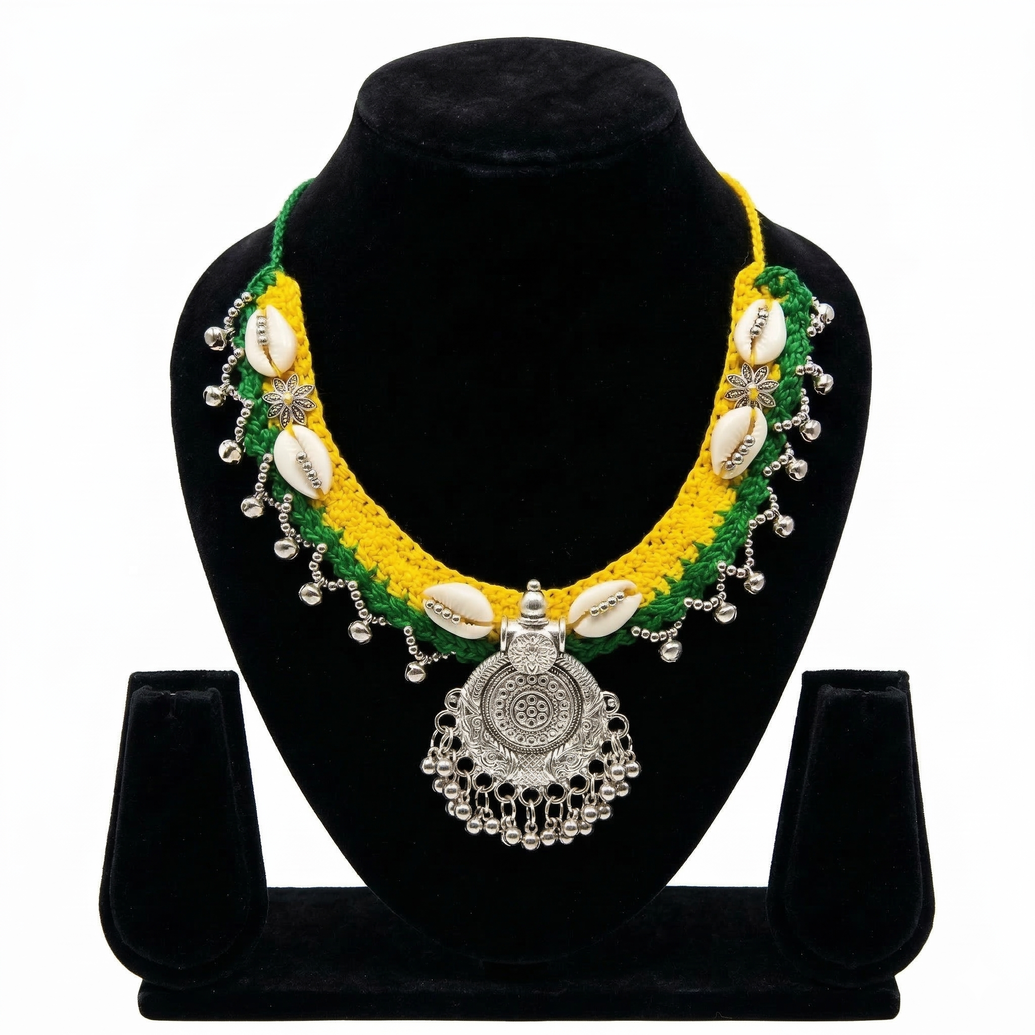 Vibrant yellow and green crochet necklace on a black bust featuring white cowrie shells, silver flower accents, and a large circular oxidized silver pendant with dangling beads.