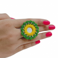 Close-up of a hand wearing a handmade green and yellow crochet flower ring featuring a central mirror and silver bead accents on a white background.