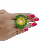 Close-up of a hand wearing a handmade green and yellow crochet flower ring featuring a central mirror and silver bead accents on a white background.