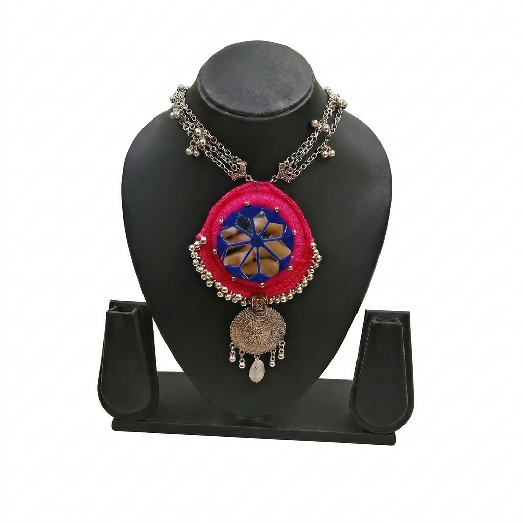 Pink handcrafted fabric necklace with blue mirror work and silver oxidized circular pendant on a black display stand.
