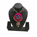 Pink handcrafted fabric necklace with blue mirror work and silver oxidized circular pendant on a black display stand.