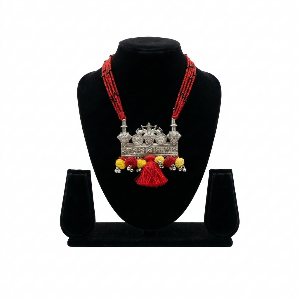 Handcrafted oxidized silver pendant necklace featuring peacock motifs, red multi-strand thread, and vibrant red and yellow pom-pom tassels on a display stand.