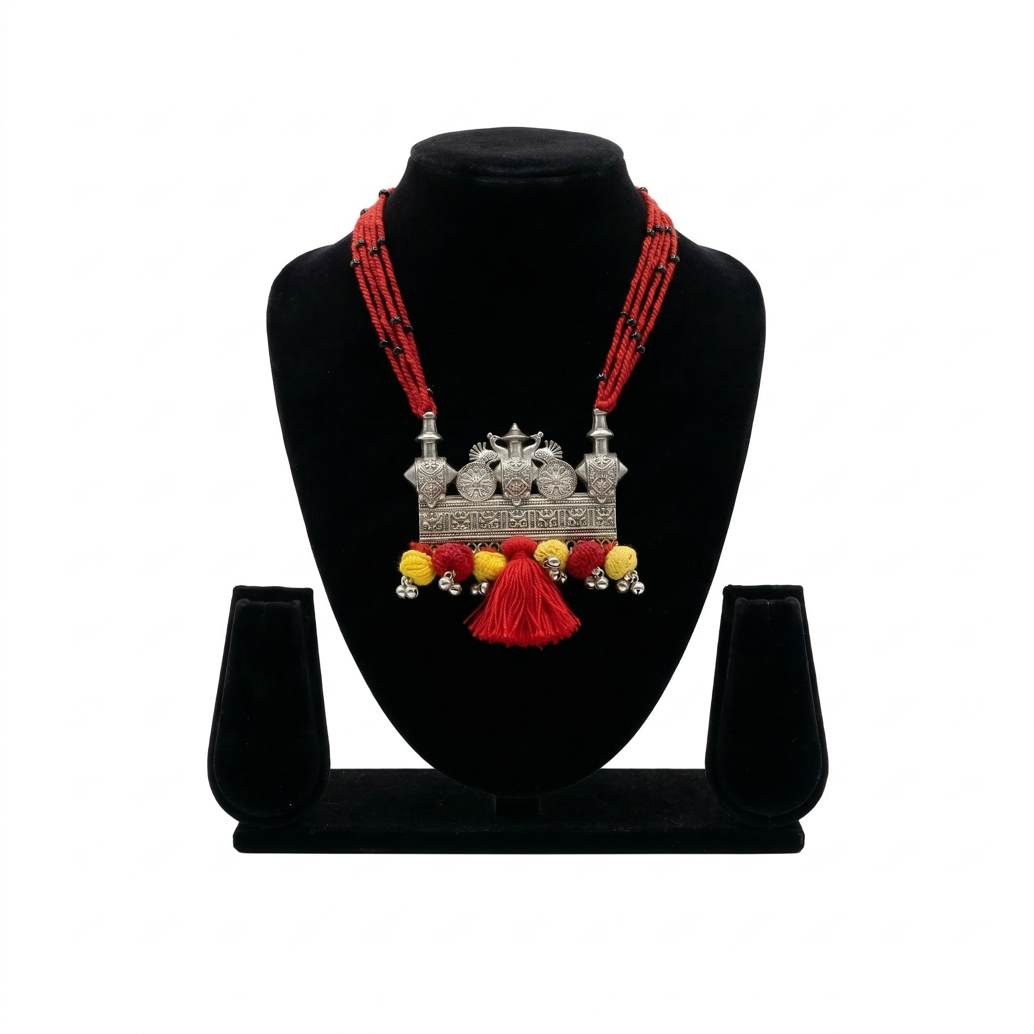 Handcrafted oxidized silver pendant necklace featuring peacock motifs, red multi-strand thread, and vibrant red and yellow pom-pom tassels on a display stand.